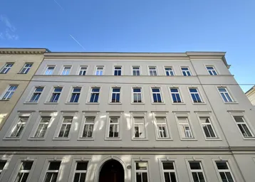 Die helle Fassade des Mehrfamilienhauses mit vielen Fenstern unter blauem Himmel.