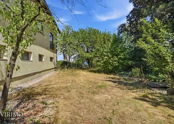 Seitenansicht des Gebäudes mit einem trockenen Gartenbereich und altem Baumbestand unter blauem Himmel.