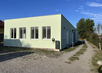 Außenansicht des Gebäudes mit hellgrüner Fassade, Fenstern und einer Ladestation für Elektrofahrzeuge.
