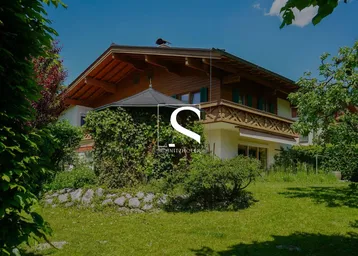 Traditionelles Holzhaus mit gepflegtem Garten und Laube unter blauem Himmel.