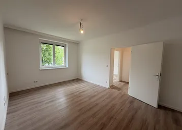 Helles, leeres Zimmer mit Laminatboden und Blick ins Grüne durch das Fenster.