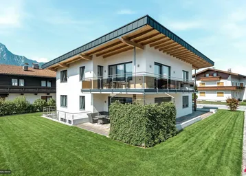 Modernes Einfamilienhaus mit weißer Fassade, großem Garten und Bergblick unter blauem Himmel.