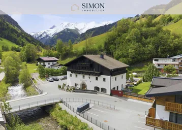 Traditionelles Gebäude in einem malerischen Tal mit schneebedeckten Bergen und üppiger grüner Landschaft.