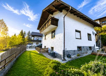 Hausfassade mit gepflegtem Garten, Rasenfläche und traditionellem Holzbalkon unter blauem Himmel.