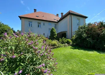 Gepflegter Garten mit weitläufiger Rasenfläche und blühenden Sträuchern vor einem traditionellen Gebäude an einem sonnigen Tag.