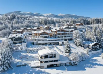 Winterlandschaft mit mehreren modernen Chalets und schneebedeckten Bergen im Hintergrund unter blauem Himmel.