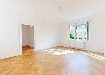 Helles Zimmer mit Fischgrätparkettboden und weißen Wänden, durchflutet von natürlichem Sonnenlicht.