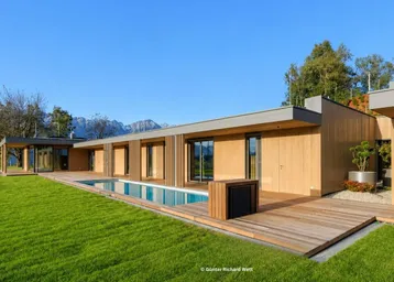 Moderne Villa mit Holzfassade, langem Pool und weitläufigem Garten vor alpiner Bergkulisse.