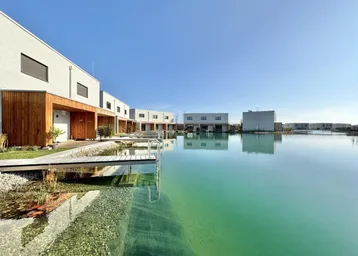 Moderne Häuser am Seeufer mit Holzfassaden und Stegen, umgeben von klarem Wasser unter blauem Himmel.