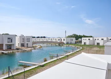 Moderne Reihenhäuser an einer künstlichen Wasserlandschaft mit Brücke unter blauem Himmel.