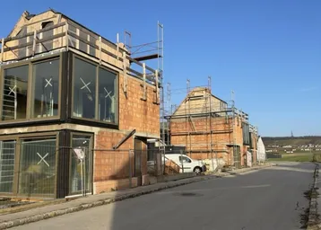 Zwei im Bau befindliche Häuser mit großen Fenstern und Gerüsten an einer ruhigen Straße.