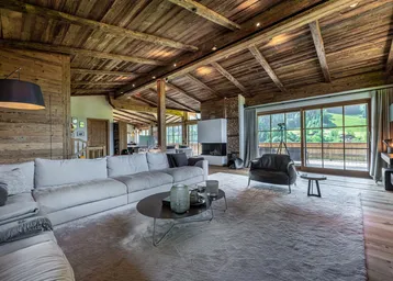 Gemütliches Wohnzimmer im Chalet-Stil mit Holzbalkendecke, Kamin und großen Fenstern mit Bergblick.