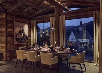 Elegantes Esszimmer mit Holzakzenten, großem Esstisch und malerischem Blick auf das Bergdorf bei Abenddämmerung.
