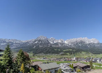 Panoramablick auf die Alpenlandschaft mit malerischem Dorf und grünen Hügeln unter blauem Himmel.