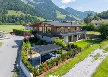 Modernes Haus mit Holzfassade, großzügigen Balkonen, Garten und Pergola mit Whirlpool vor Bergkulisse.