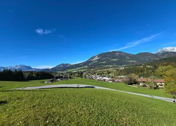 Weitläufige grüne Hügellandschaft mit einem Dorf im Tal und majestätischen Bergen im Hintergrund unter blauem Himmel.