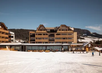 Modernes Hotelgebäude in den Bergen mit Skihang im Vordergrund und sonnigem Wetter.