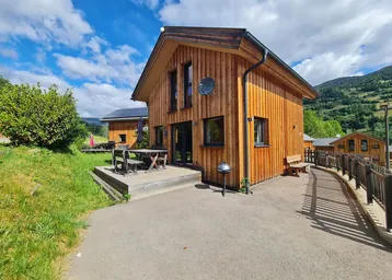 Modernes Holzhaus mit Terrasse und Bergblick unter blauem Himmel und viel Grün.
