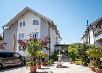 Elegantes Gebäude mit gepflegtem Eingangsbereich, Springbrunnen und Palmen in Töpfen unter blauem Himmel.