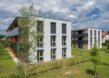 Moderne Mehrfamilienhäuser mit weißer Fassade, dunklen Balkonen und grüner Umgebung unter bewölktem Himmel.