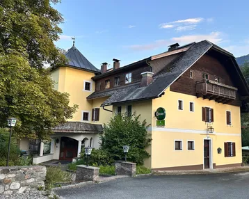 Historisches Gebäude mit gelber Fassade, dunklem Dach und einem markanten Turm, umgeben von Bäumen.