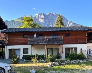 Modernes Haus mit Holzfassade und Balkon, umgeben von Grünflächen mit beeindruckendem Bergblick.