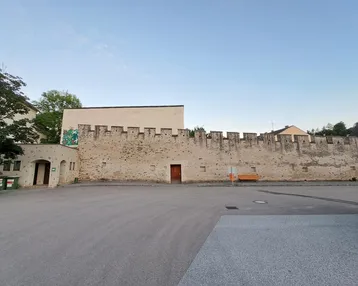 Alte Stadtmauer mit Zinnen und Holztür, umgeben von einem gepflasterten Platz.