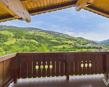 Traditioneller Holzbalkon mit geschnitztem Geländer und weitem Blick auf die grünen Berge und ein Dorf.