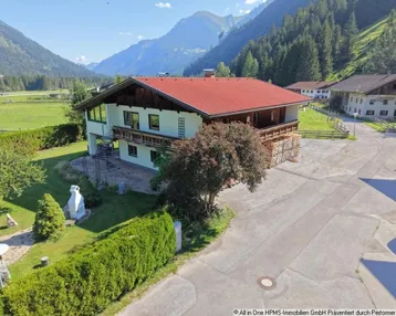 Traditionelles Haus mit rotem Dach und Holzbalkonen, umgeben von grüner Landschaft und Bergen.