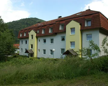 Mehrfamilienhaus mit Mansarddach, hellgrauer Fassade und gelben Akzenten, umgeben von grüner Natur und Hügeln.