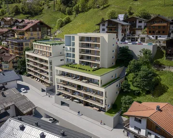 Moderne Apartmentgebäude mit begrünten Dächern und Balkonen in alpiner Umgebung, umgeben von grünen Hügeln.
