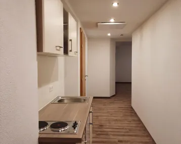 Schmaler Flur mit integrierter Küchenzeile, Laminatboden und weißen Wänden, beleuchtet durch Deckenspots.