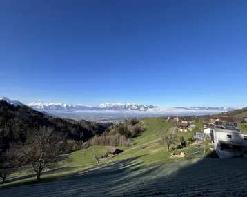 Panoramablick über ein grünes Tal mit einer Stadt in der Ferne und schneebedeckten Bergen unter blauem Himmel.