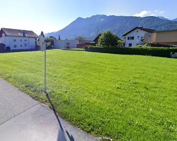 Weitläufige Grünfläche mit Bergblick und umliegenden Wohnhäusern unter blauem Himmel.