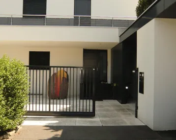Moderne Hausfassade mit Eingangsbereich, schwarzem Zaun und Kunstobjekt.