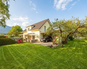 Haus mit großem Garten, Terrasse und Carport unter blauem Himmel und Bergblick.