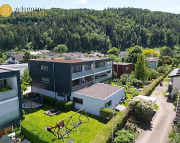 Moderne Wohnanlage mit Flachdach, Balkonen und einem gepflegten Garten mit Spielgeräten, umgeben von viel Grün.