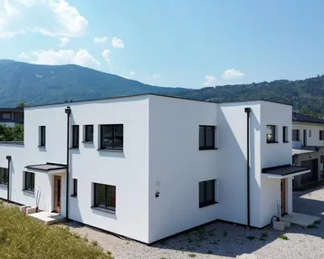 Modernes weißes Doppelhaus mit Flachdach und Bergblick unter blauem Himmel.
