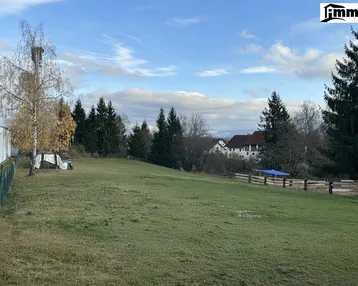 Weitläufiges grünes Grundstück mit Bäumen und einem Haus im Hintergrund unter bewölktem Himmel.