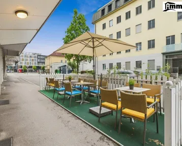 Außenbereich mit Tischen, Stühlen und Sonnenschirm, umgeben von einem weißen Zaun.
