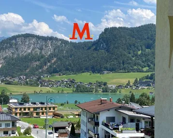 Panoramablick auf den See und die umliegenden Berge unter blauem Himmel mit leichter Bewölkung.