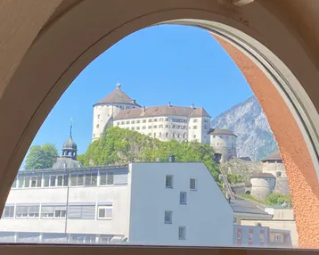 Malerischer Blick durch ein Bogenfenster auf eine historische Burg auf einem Hügel und Berge.
