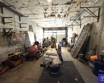 Großer, unaufgeräumter Werkstatt- oder Lagerraum mit Betonwänden, offenem Deckenbereich und verschiedenen Geräten.
