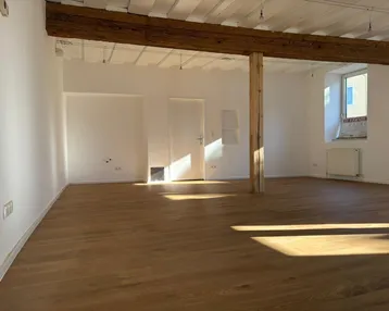 Geräumiger Innenraum mit Holzbalken, Holzboden und viel natürlichem Licht durch die Fenster.