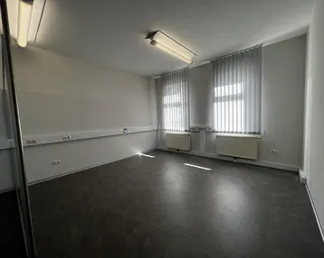 Leerer Büroraum mit dunklem Boden, hellen Wänden und vertikalen Jalousien an den Fenstern.