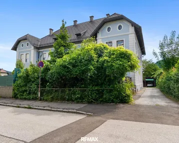 Historisches Gebäude mit gepflegter Fassade und üppiger Bepflanzung an der Straße.