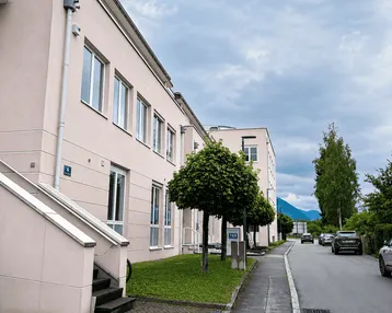 Gebäudeansicht von außen mit hellen Fassaden, Fenstern und einem Gehweg entlang der Straße unter bewölktem Himmel.