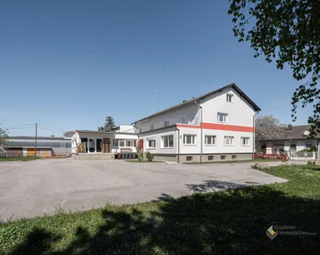 Gebäudeansicht mit großem Vorplatz und umliegender Grünfläche unter blauem Himmel.