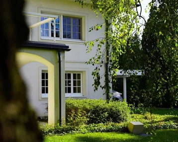 Teilansicht des Hauses mit gepflegtem Vorgarten und üppigem Grün an einem sonnigen Tag.