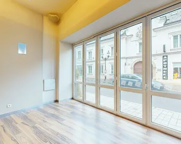 Heller Innenraum mit großen Fenstern zur Straße, Holzboden und moderner Deckenbeleuchtung.